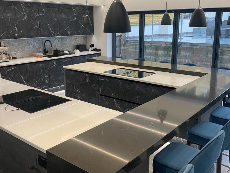A finished kitchen by Colebarn Interior Design, Derbyshire. The kitchen is modern with lights hanging from the ceiling. The walls are painted in a light grey colour with chrome work tops and marbled fascias.
